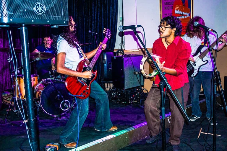Adelaide band Jaded Earth performing live on stage with electric guitars, vocals, tambourine, and drums at a venue.